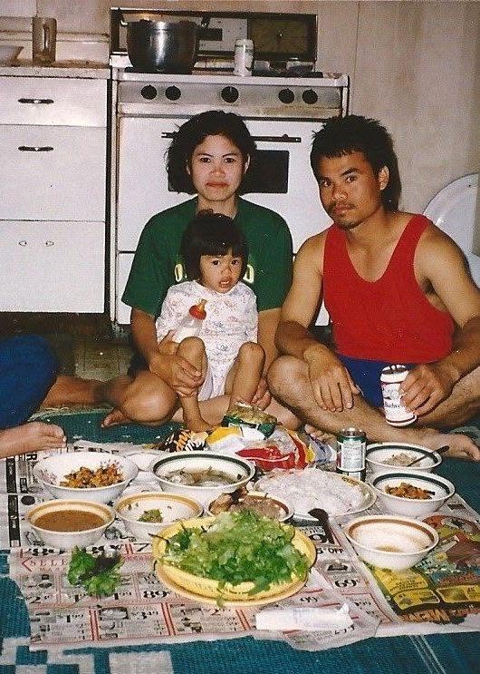Me as a toddler on my mom's lap next to my dad, a Lao dinner spread out on newspaper on the kitchen floor